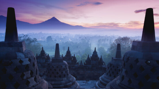 Stone stupas of an ancient temple overlooking a misty landscape and mountain at dawn.