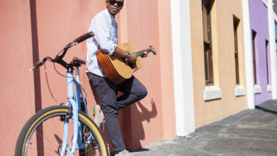 A man with a guitar and a blue bicycle standing against a bright pink building.