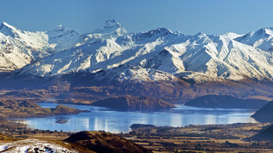 Panoramic view of jagged snow-capped mountain peaks above a blue lake and a rural settlement.