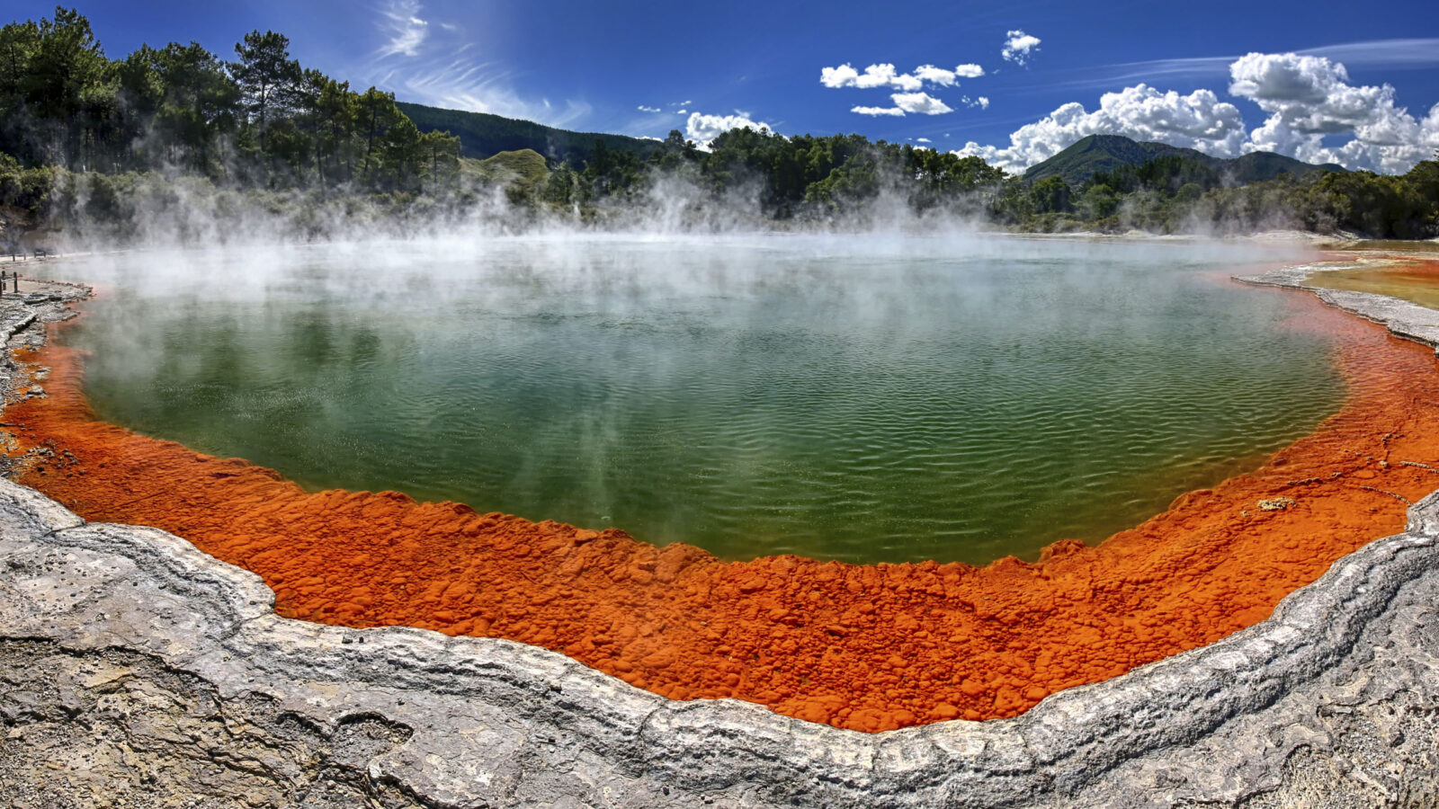 A steaming geothermal spring with a bright orange rim and green-tinted water.