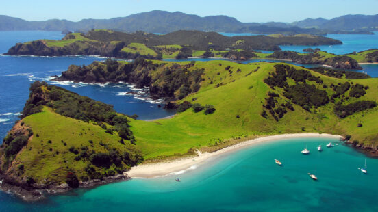 Aerial view of green hilly islands and a crescent beach with boats anchored in clear blue water.