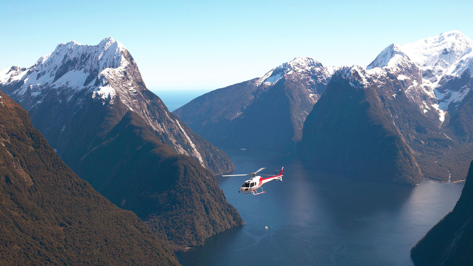 A red and white helicopter soars through Milford Sound, framed by dramatic, snow-capped peaks and deep fiord waters.