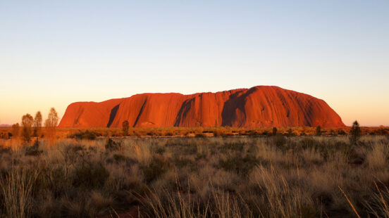 A large red sandstone monolith under a clear sky in a desert landscape.