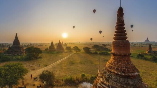 Multiple hot air balloons float over the ancient temples and pagodas of Bagan, Myanmar, during a hazy golden sunrise.