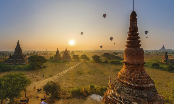 Multiple hot air balloons float over the ancient temples and pagodas of Bagan, Myanmar, during a hazy golden sunrise.