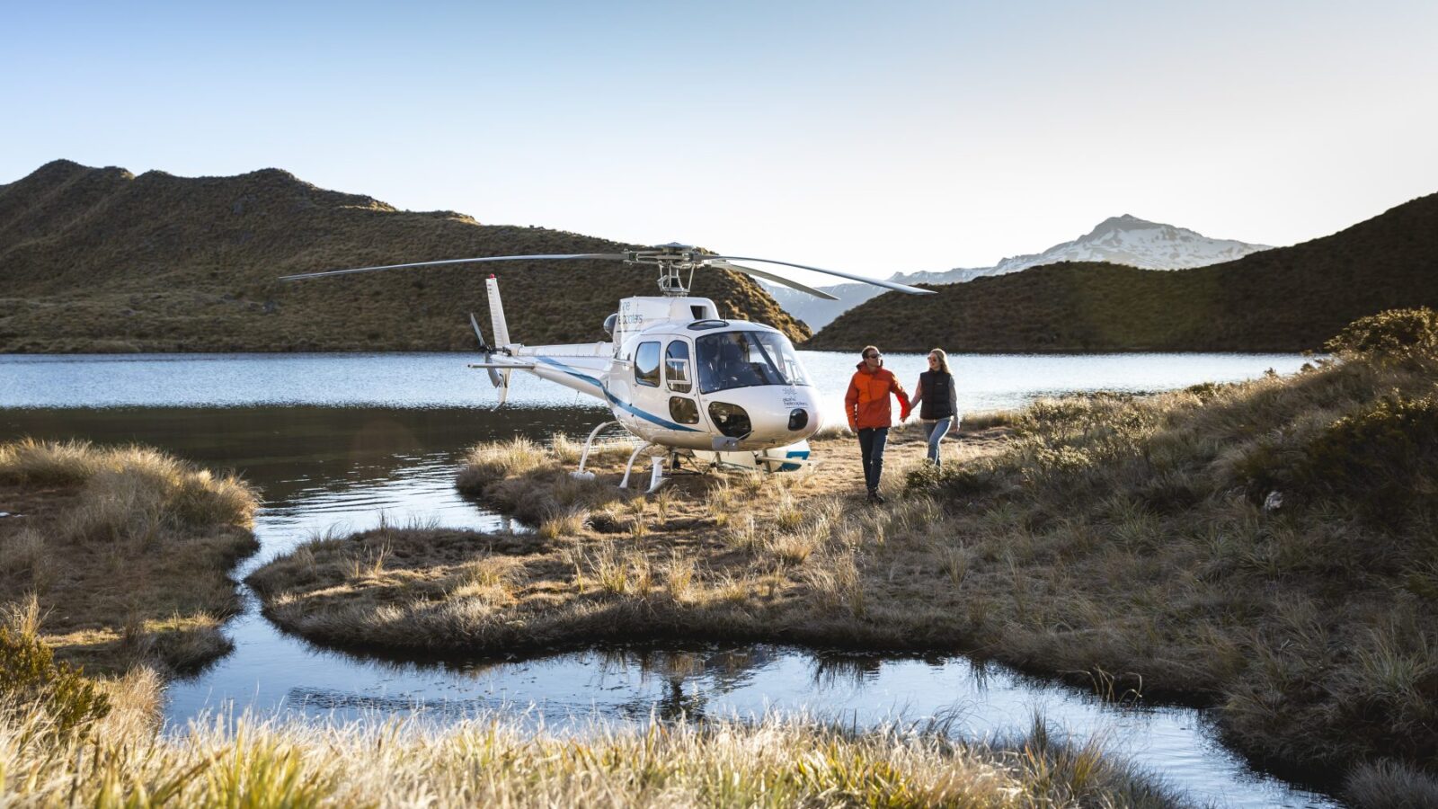 A couple holds hands near a white helicopter parked beside a remote alpine lake in New Zealand.