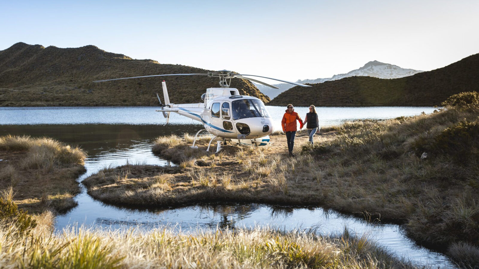 Two people standing by a helicopter that has landed in a scenic plain near a lake