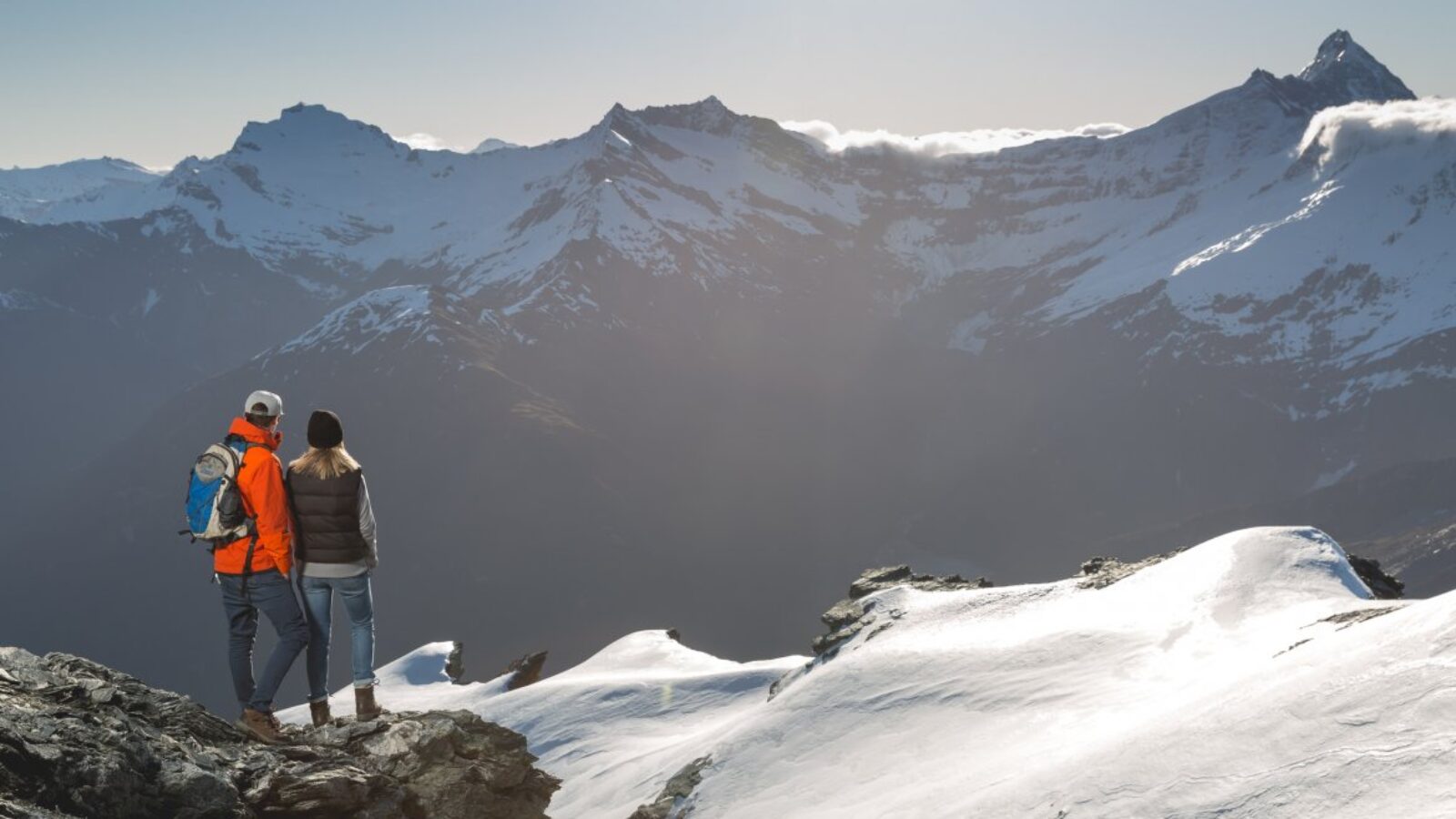 Two hikers take in panoramic views of jagged, snow-dusted peaks from a rocky mountain ridge in New Zealand.