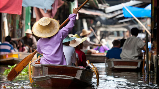 People wearing traditional hats row wooden boats through a busy floating market canal.