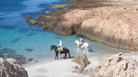 Two people riding a black horse and a white horse on a sandy beach by clear turquoise water and rocky hills.