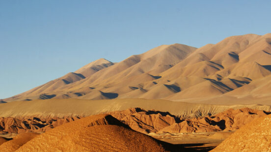 Smooth, large sand dunes rise above rugged red terrain under a clear blue sky in the Puna.