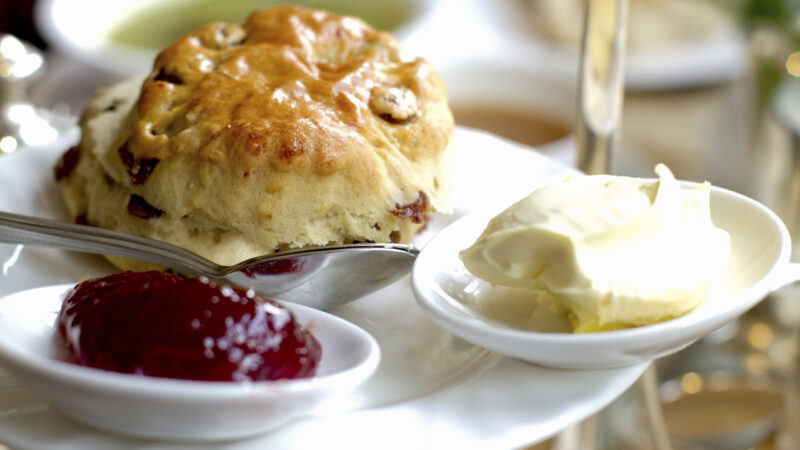 A golden-brown fruit scone served with a side of red strawberry jam and thick clotted cream on white chinaware.