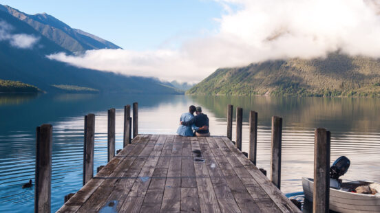 A couple embraces at the end of a wooden pier, taking in tranquil, mist-shrouded mountain views over Lake Rotoiti in Nelson Lakes National Park, New Zealand.