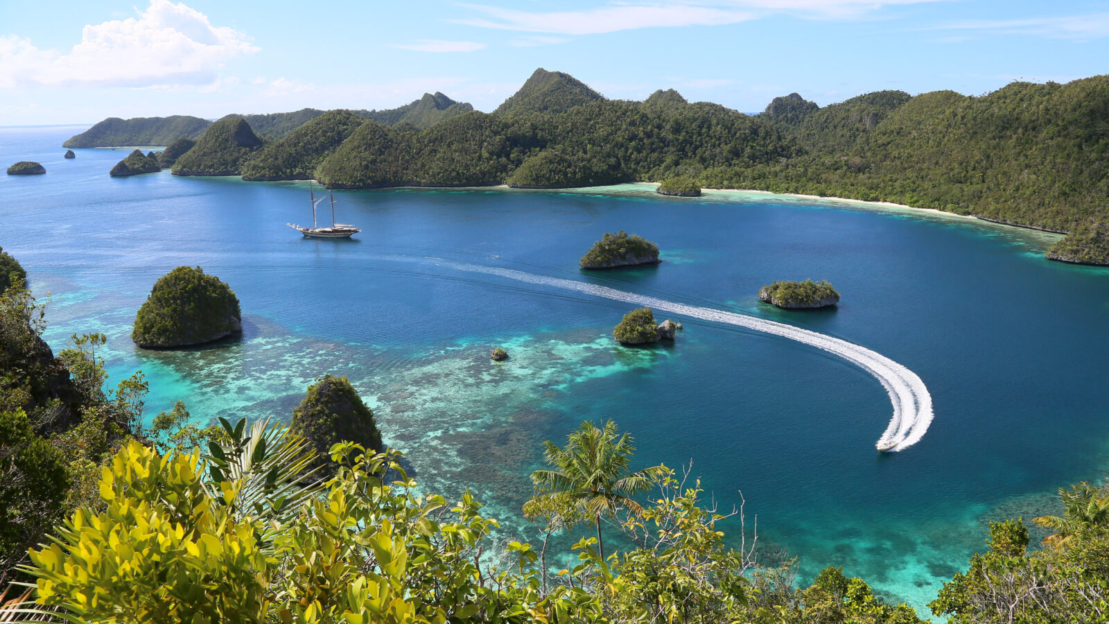 A speedboat leaves a white wake through a clear blue lagoon surrounded by lush green islands for Asia honeymoons.