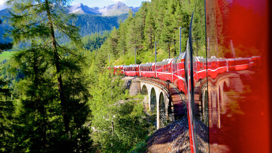 A red passenger train crossing a curved stone bridge through a coniferous forest.