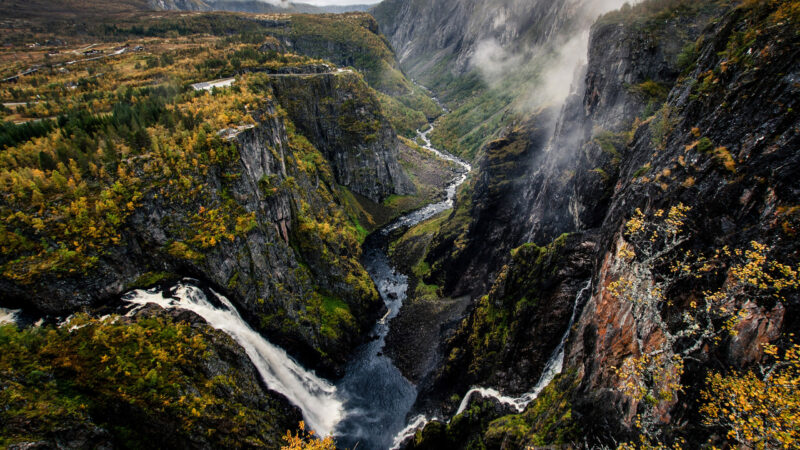 A dramatic deep canyon with a winding river and waterfall surrounded by steep, mossy rock walls and mist.