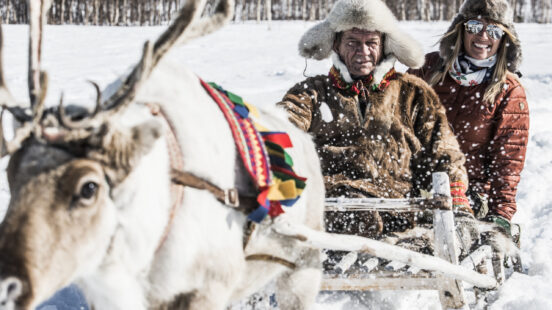 A man in a fur coat and a woman in a red jacket sitting in a reindeer-drawn sled on a sunny snowy day.
