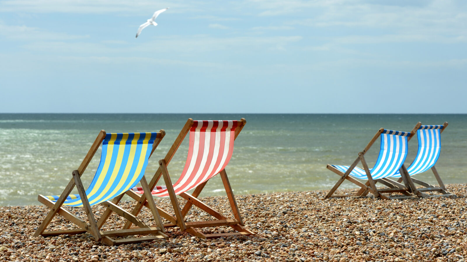 Yellow, red, and blue striped beach chairs on a shingle shore overlooking the ocean with a bird in flight.