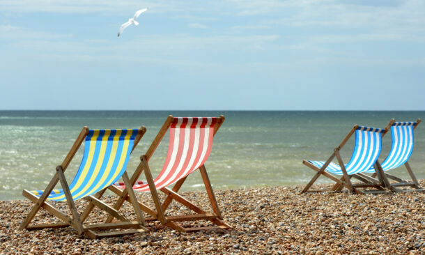 Yellow, red, and blue striped beach chairs on a shingle shore overlooking the ocean with a bird in flight.