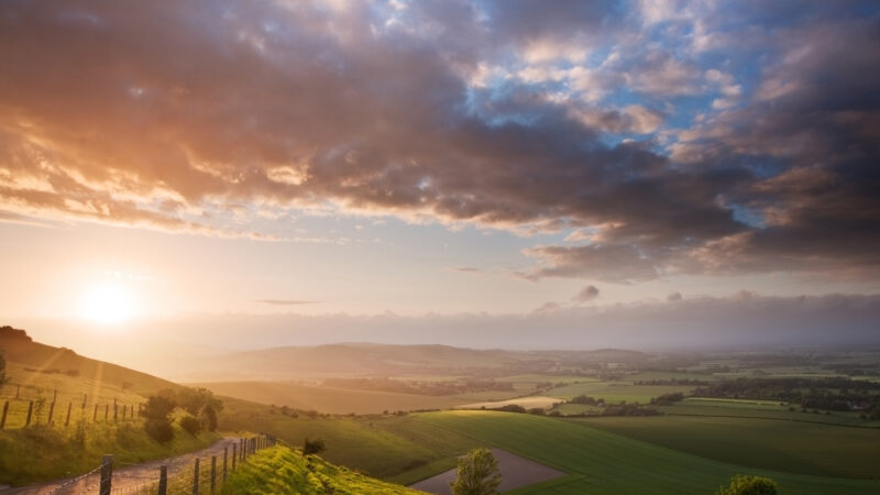 A bright sunset over a lush valley of green fields and rolling hills under a sky filled with textured clouds.