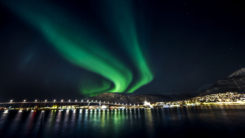 Bright green northern lights waving in the dark sky over a glowing city waterfront and long bridge.