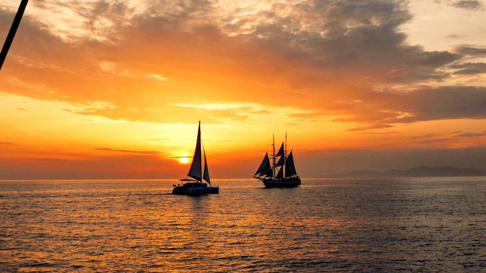 Silhouette of two sailboats on the sea during a vibrant orange and yellow sunset with scattered clouds.