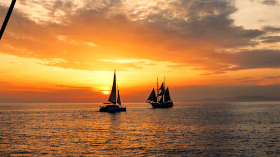 Silhouette of two sailboats on the sea during a vibrant orange and yellow sunset with scattered clouds.