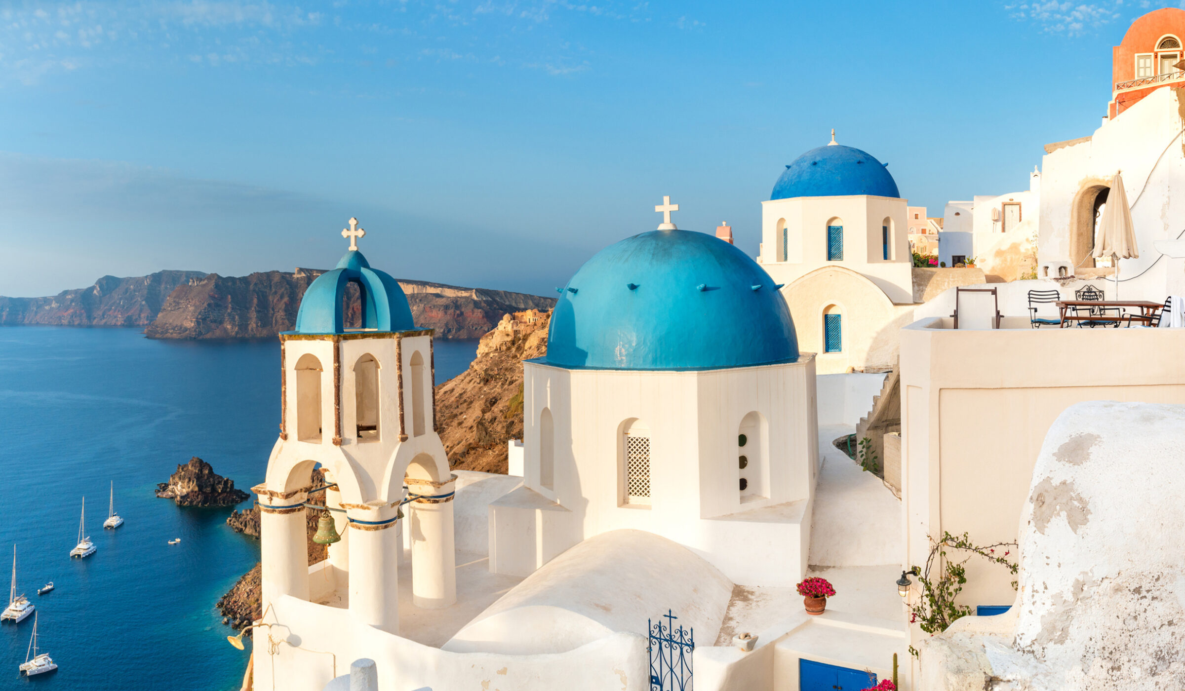 White churches with bright blue domes on a cliffside overlooking a deep blue sea under a clear sky.