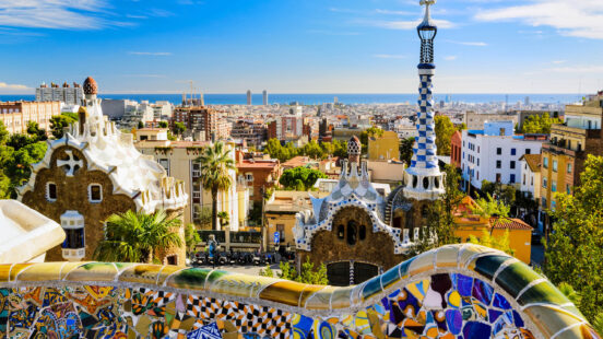 View of Park Guellx27;s mosaic walls and iconic buildings with the Barcelona city skyline in the background.