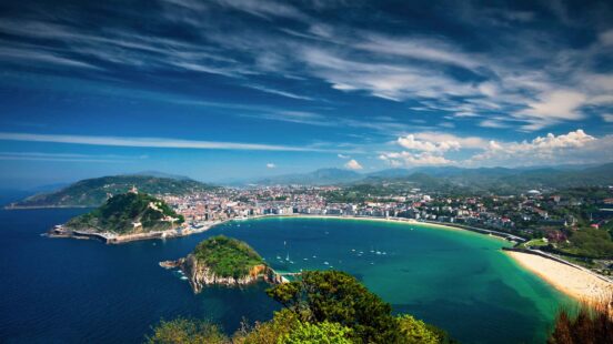 High angle view of a turquoise bay and sandy beach surrounded by lush green mountains and a coastal city.