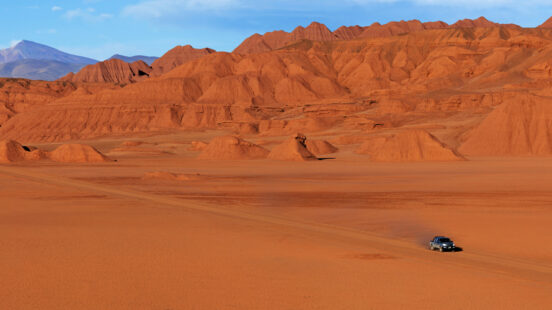 A vehicle driving through a wide desert valley surrounded by jagged red mountains.