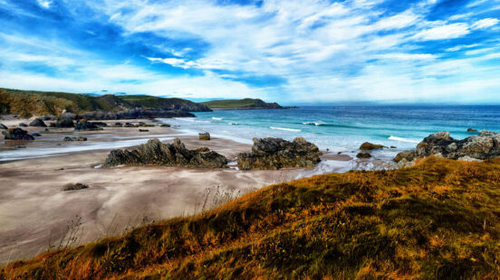 A scenic coastline with jagged rocks on a sandy beach, turquoise ocean water, and a vibrant blue sky with clouds.