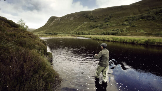 A man fly fishing in a river surrounded by rolling green hills under an overcast sky.