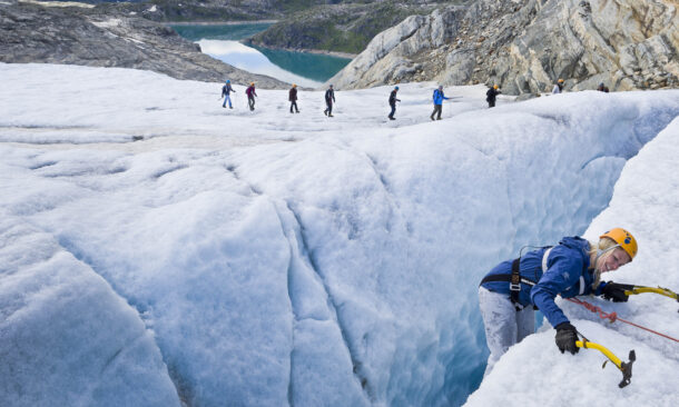 A person ice climbing in a glacier crevasse while others walk across the ice in the background.