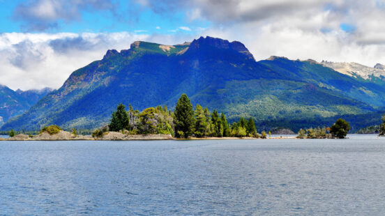 A deep blue lake with a small forested island in the foreground and a large, dark mountain in the background.