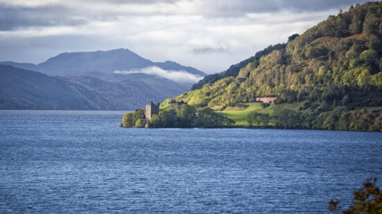 Stone ruins of Urquhart Castle on the shore of Loch Ness with mountains in the background.