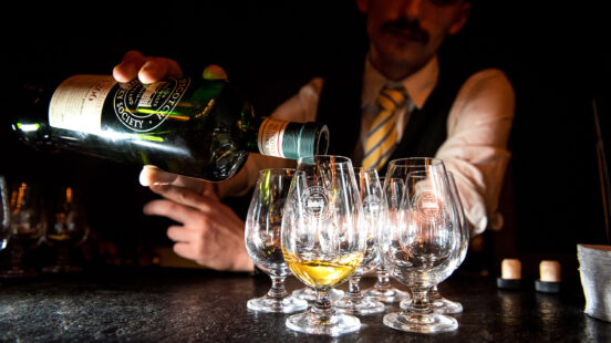 Close-up of a hand pouring amber liquid from a whisky bottle into several specialized tasting glasses.