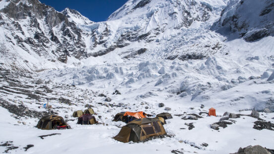 Several expedition tents are pitched on a snowy slope at the foot of a massive, jagged glacier. The sky is clear blue.