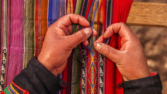 Close-up of two hands weaving intricate blue and red threads into a bright, traditional Peruvian textile on a loom.