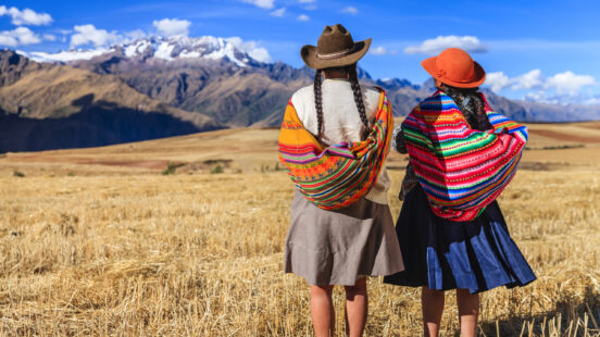 Two people in colorful wraps and hats stand in a field facing distant snowy mountains.