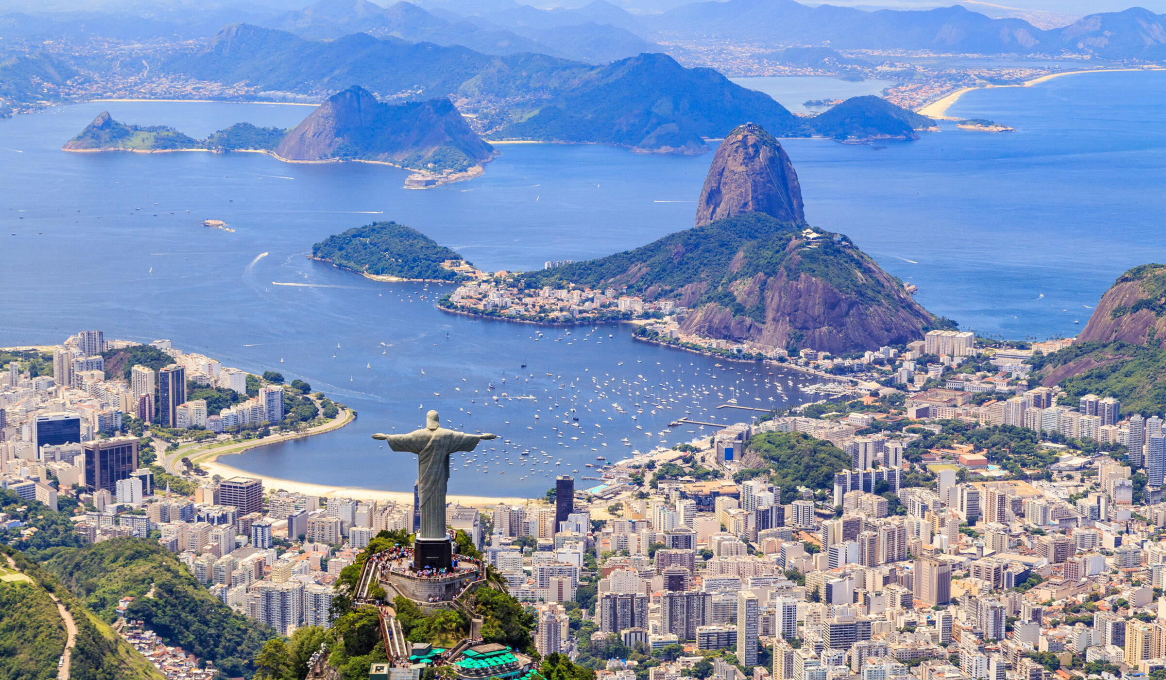 The Christ the Redeemer statue stands on a mountain peak overlooking a coastal city and bay.