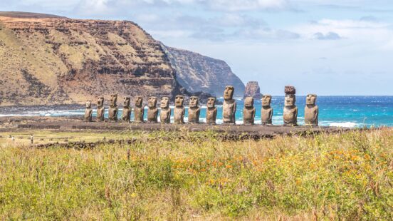 A row of Moai stone statues on Easter Island facing away from the blue ocean and tall cliffs.