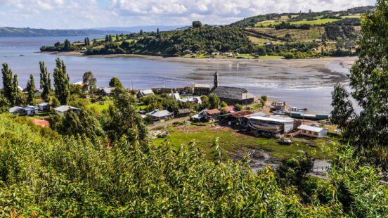 A small coastal village with a dark church is nestled in a green valley by the water, surrounded by hills.