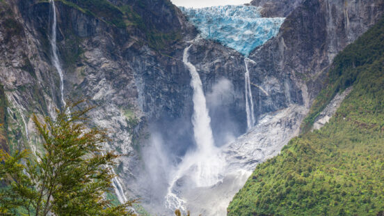 Multiple waterfalls cascade down a rocky cliff face beneath the blue ice of a glacier, surrounded by green trees.