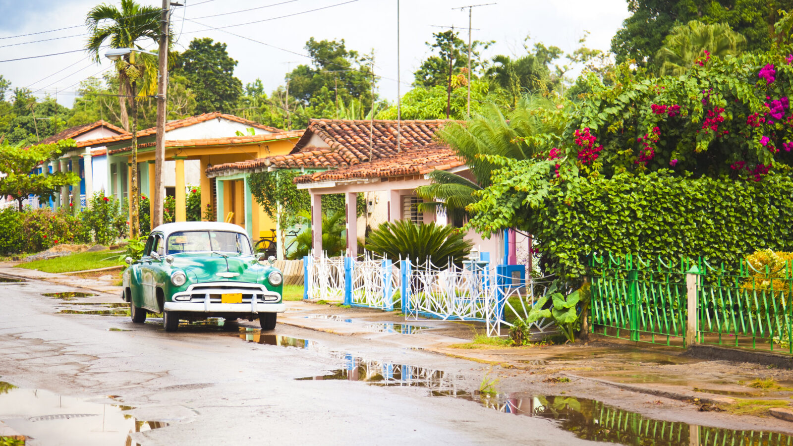 A vintage green and white car parked on a residential street with puddles and colorful houses.