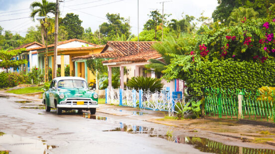 A vintage green and white car parked on a residential street with puddles and colorful houses.
