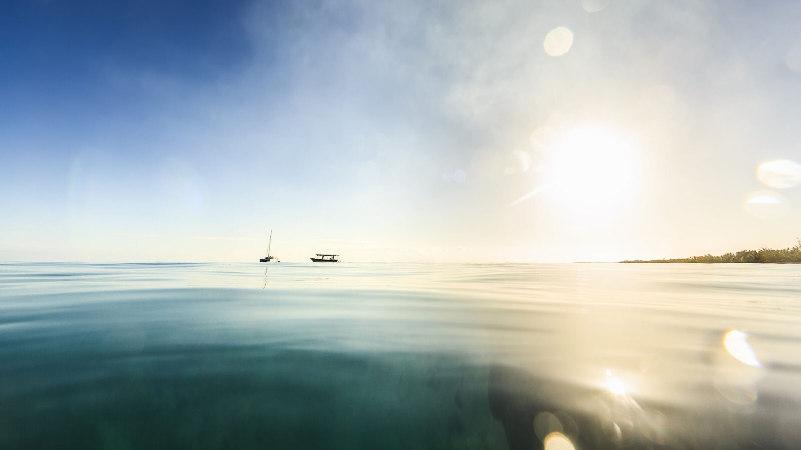 A DSLR photo taken from inside the sea, just above surface level, in tranquil water in Rangiroa, French Polynesia. The sun is low on the horizon and will bet set in a few moments. It is produncing some flare in the water drops on the lens. Two boats are anchored to the left of the frame.