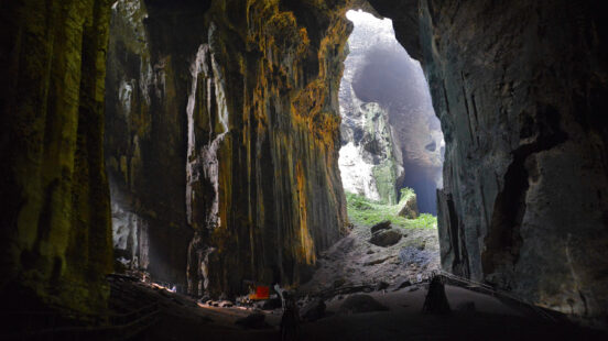 Perspective from inside a giant cave looking toward a bright opening with tall, textured rock formations.