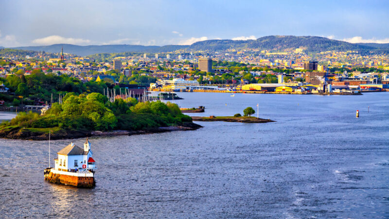A small white lighthouse building on a rocky outcrop in the water with a city and green hills in the background.