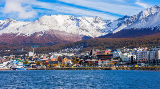 A cityscape of Ushuaia, Argentina, on the coast with colorful buildings, set against large snow-covered mountains in the background.